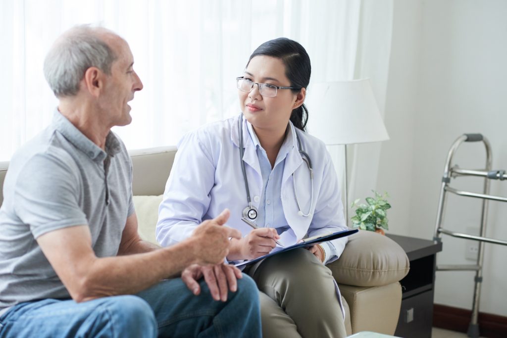 Asian adult woman with clipboard sitting on sofa in house of senior man during visit and listening to patient