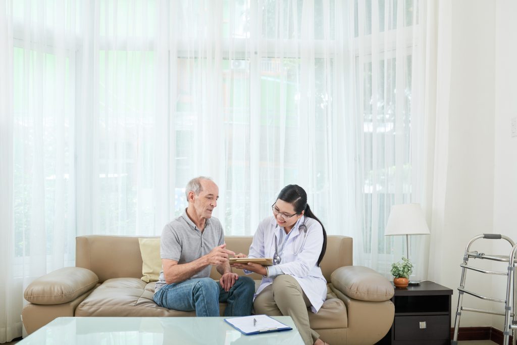 Adult Asian woman showing tablet to elderly man sitting on sofa at home during visit